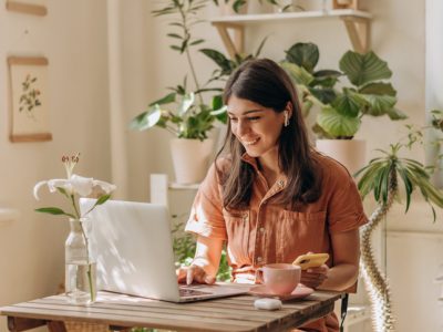 Positive young mixed race woman using a laptop and smartphone at home.Cozy home interior with indoor plants.Remote work, business,freelance,online shopping,e-learning,urban jungle concept