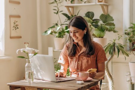 Positive young mixed race woman using a laptop and smartphone at home.Cozy home interior with indoor plants.Remote work, business,freelance,online shopping,e-learning,urban jungle concept Positive young mixed race woman using a laptop and smartphone at home.Cozy home interior with indoor plants.Remote work, business,freelance,online shopping,e-learning,urban jungle concept