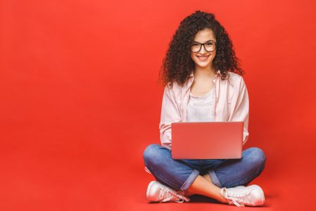 Happy young curly beautiful woman sitting on the floor with crossed legs and using laptop on red background. Happy young curly beautiful woman sitting on the floor with crossed legs and using laptop on red background.