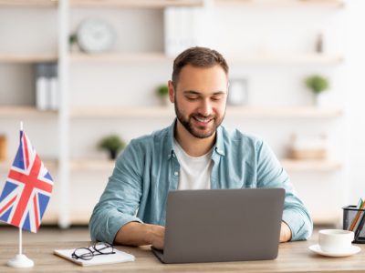 Happy young guy with flag of Great Britain working at desk with laptop in home office