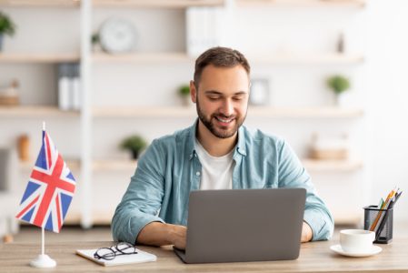 Happy young guy with flag of Great Britain working at desk with laptop in home office Happy young guy with flag of Great Britain working at desk with laptop in home office