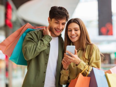 Couple Shopping Using Phone Application Holding Shopper Bags In Mall