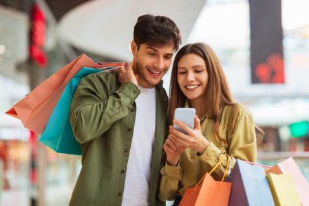 Couple Shopping Using Phone Application Holding Shopper Bags In Mall Couple Shopping Using Phone Application Holding Shopper Bags In Mall