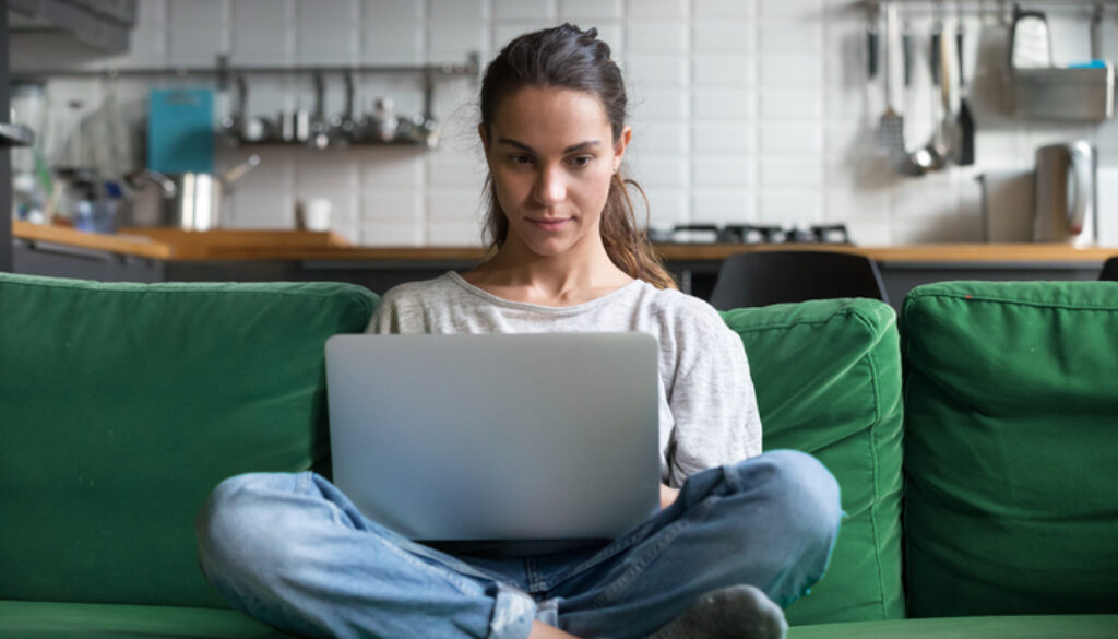 Serious woman using laptop checking news online sitting on sofa