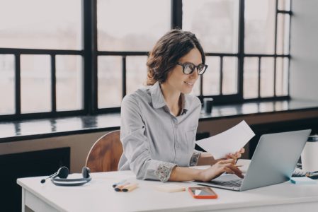 Focused European business woman sitting at home office holds financial documents, working on laptop Focused European business woman sitting at home office holds financial documents, working on laptop